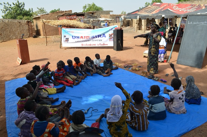 Des enfants assis en cercle sur une bâche lèvent la main pendant une séance de lecture en plein air.