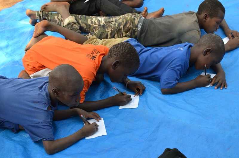 Des enfants allongés sur une bâche bleue écrivent sur des feuilles.