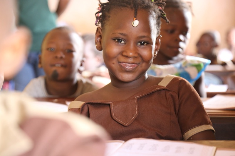 Une élève souriante assise à son pupitre, un livre ouvert devant elle, au milieu de ses camarades.